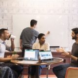 Vier collega’s overleggen rond laptops en een whiteboard op kantoor